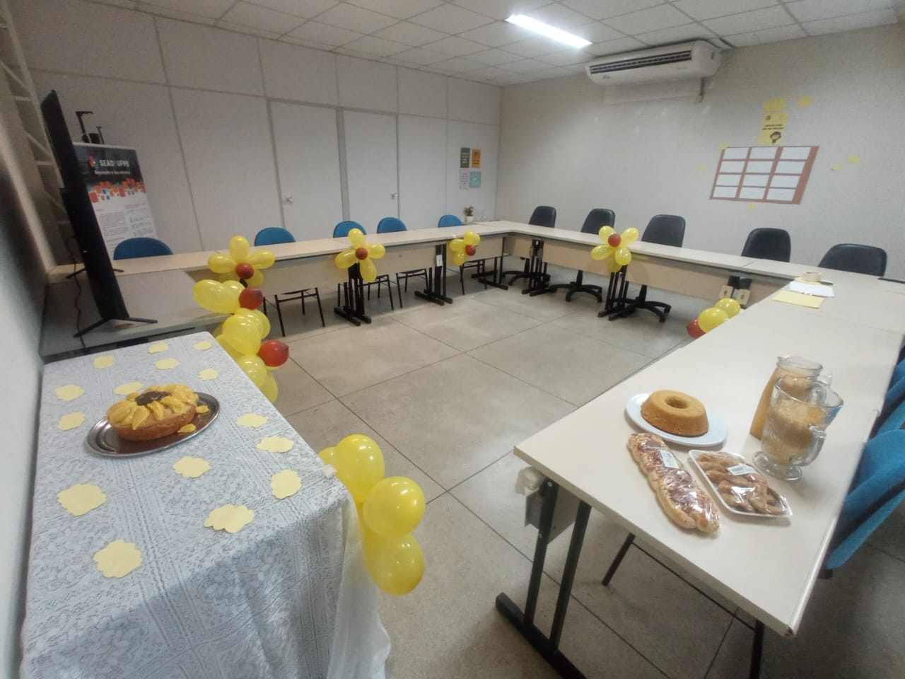 Esta é uma fotografia de uma sala de reunião decorada. À esquerda, há uma mesa coberta com uma toalha branca de renda, sobre a qual está um prato de sobremesa com fatias de bolo de cor amarela. No fundo da sala, há uma mesa em formato de